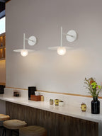 Two white Clérachic wall lights glow above a wooden bar counter set with a flower vase.