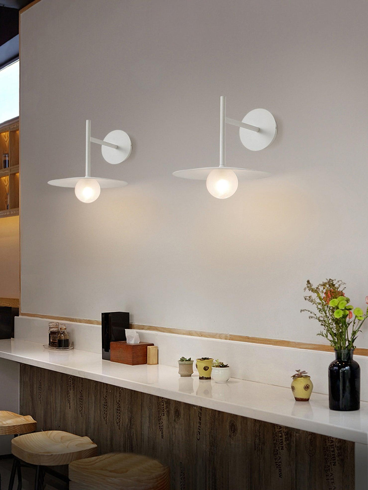 Two white Clérachic wall lights glow above a wooden bar counter set with a flower vase.