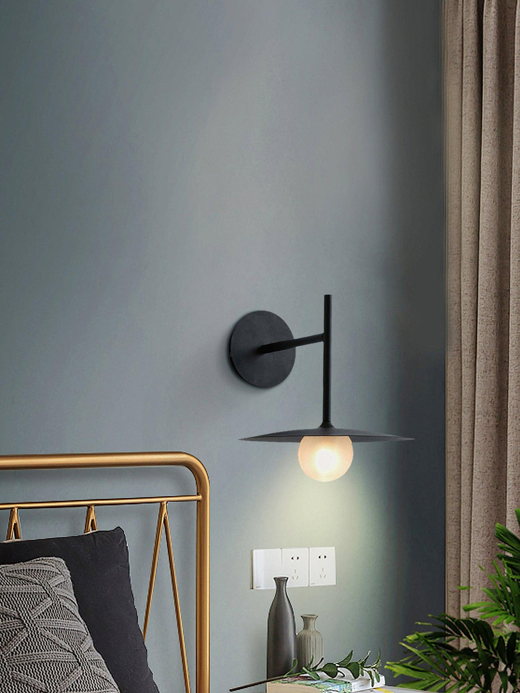 Mounted on a blue-grey wall, this black Clérachic wall light glows above a brass-framed bed.