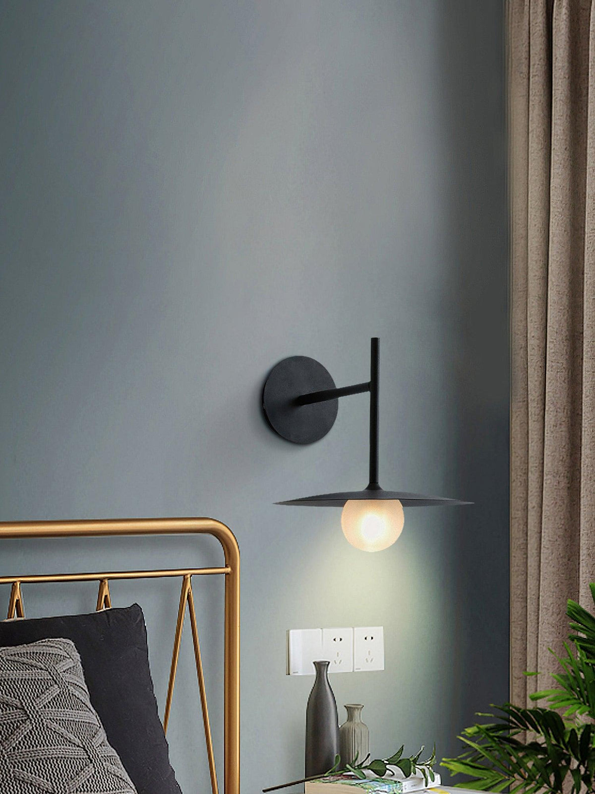 Mounted on a blue-grey wall, this black Clérachic wall light glows above a brass-framed bed.