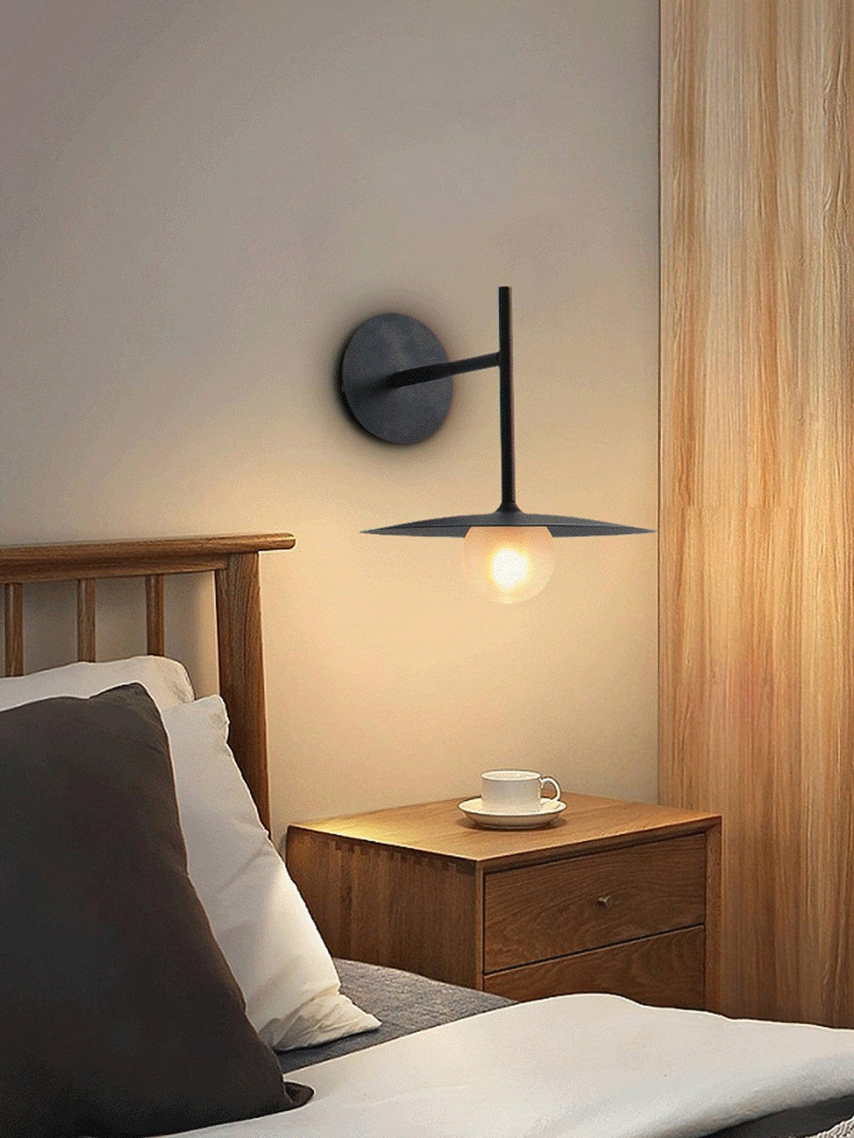 A black Clérachic wall light glows above a wood-framed bed dressed in linen pillows.