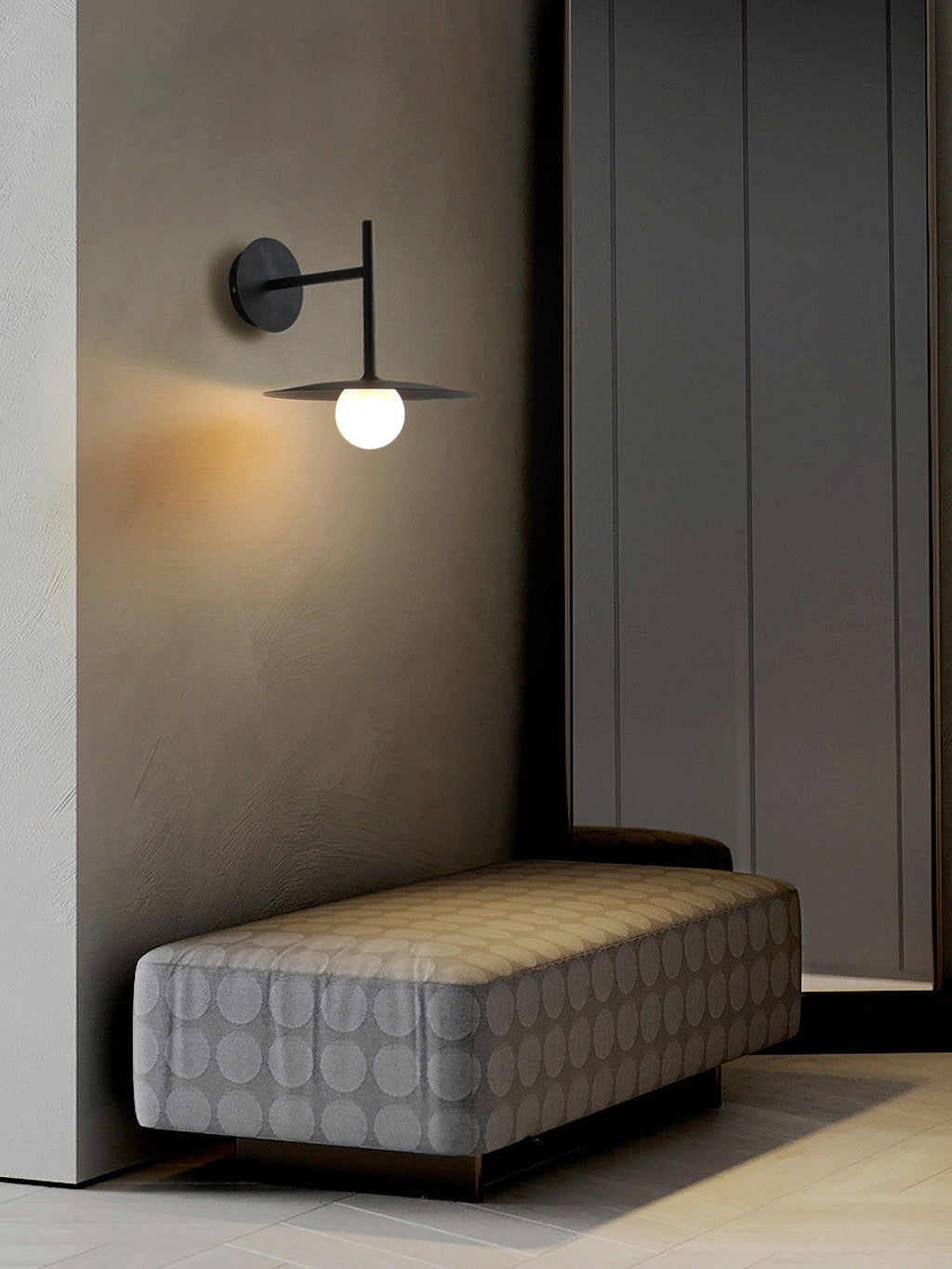 In a hallway corner, this black Clérachic wall light glows beside a tufted grey ottoman.