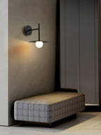 In a hallway corner, this black Clérachic wall light glows beside a tufted grey ottoman.