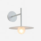 Studio view of this white Clérachic wall light, its disc shade glowing warmly.