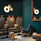 Grouped Clérachic wall lights glow on a teal wall beside a round mirror, in a lounge with leather chairs.