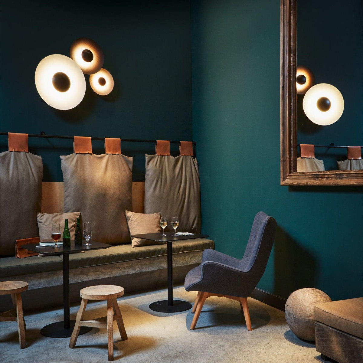 Grouped Clérachic wall lights glow on a teal wall beside a round mirror, in a lounge with leather chairs.