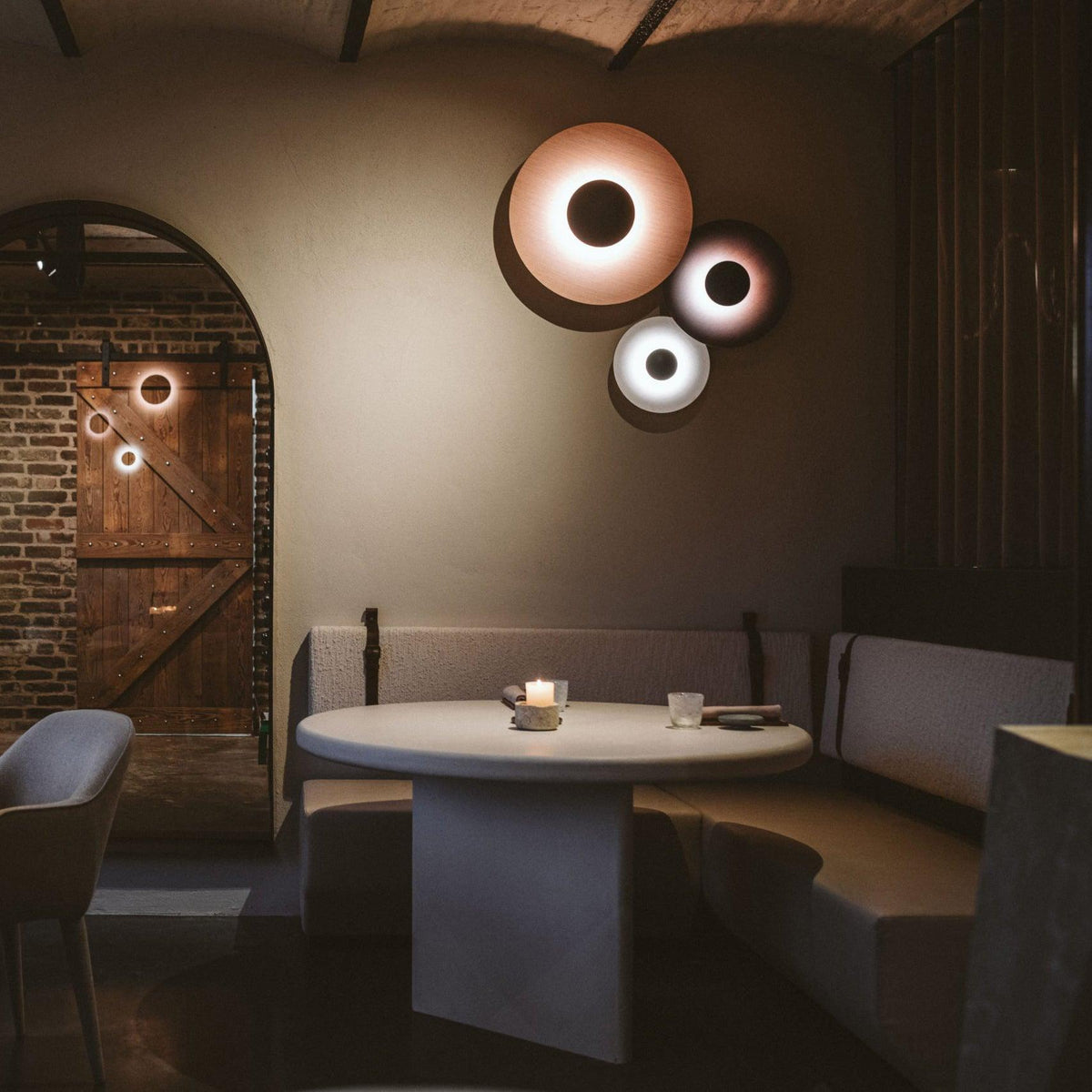 Three of this Clérachic wall light glow warm on a wall near a brick archway, above a round dining table.