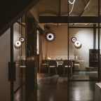 Grouped Clérachic wall lights glow near a glass-paned door in a minimalist restaurant with dining tables.