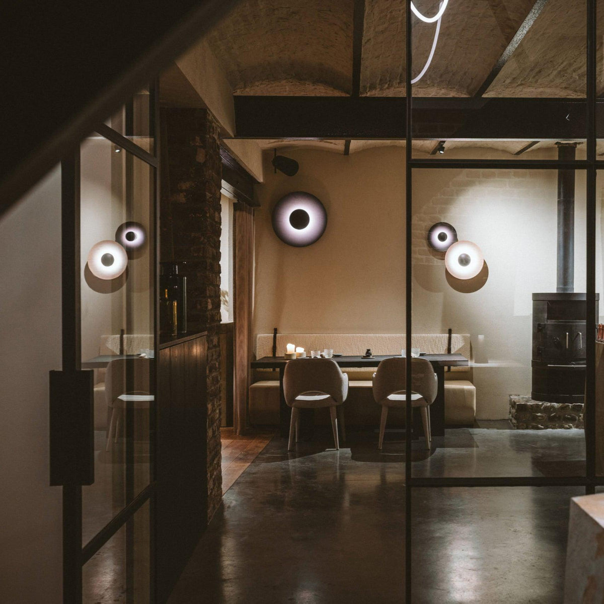 Grouped Clérachic wall lights glow near a glass-paned door in a minimalist restaurant with dining tables.
