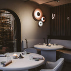 Grouped Clérachic wall lights glow warm on a textured wall near an arched doorway in a restaurant with candlelit tables.