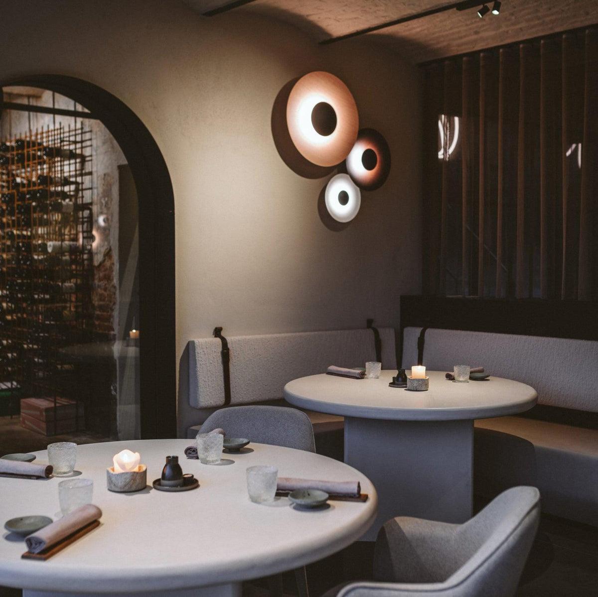 Grouped Clérachic wall lights glow warm on a textured wall near an arched doorway in a restaurant with candlelit tables.