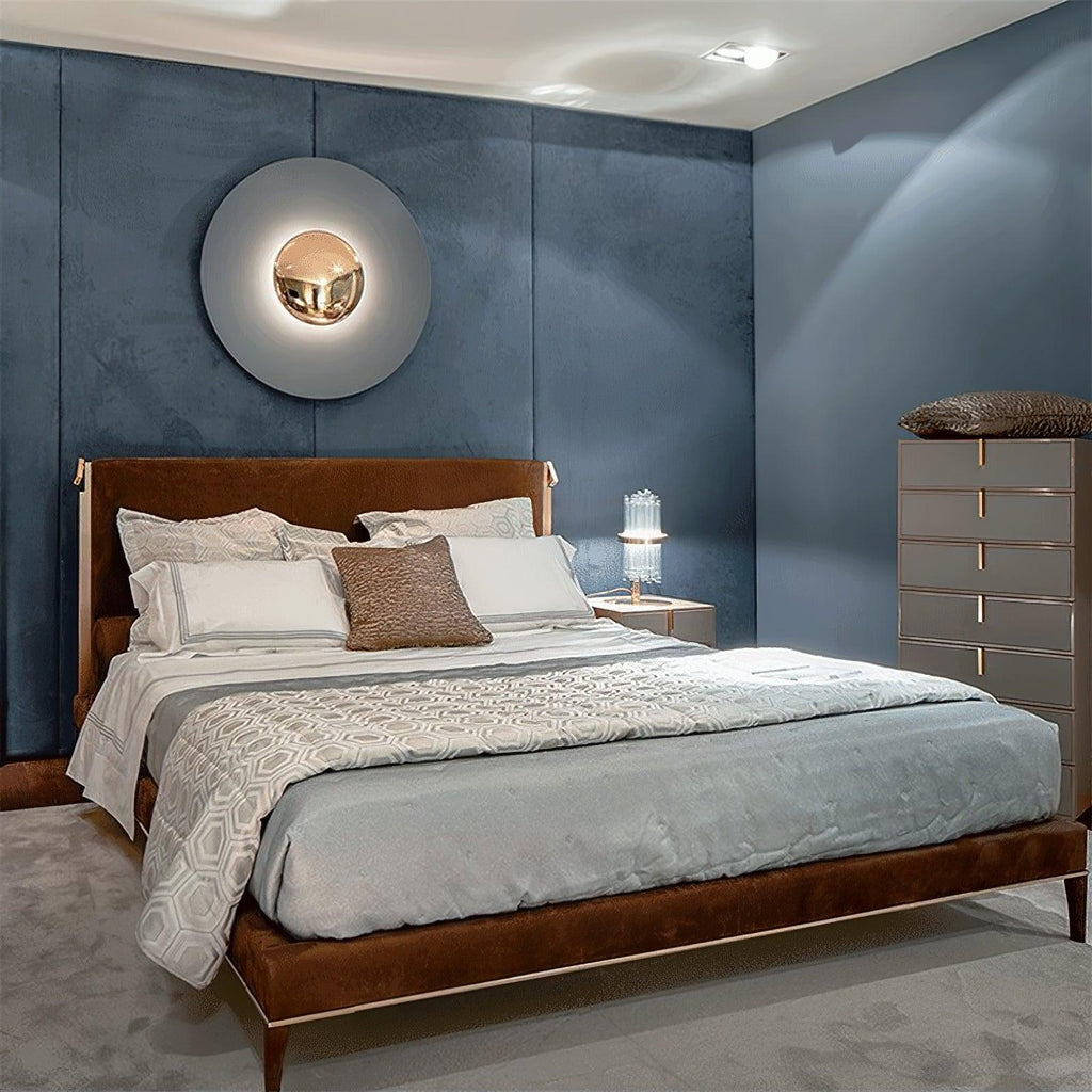 A pale grey Clérachic wall light glows above a bed with a brown leather frame, on a slate-blue wall.