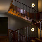 Two small Clérachic wall lights glow warm along a wood-railed staircase, seen from a landing above.