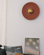 Another view of the unlit terracotta Clérachic wall light on a plain wall, beside a cushion and framed print.