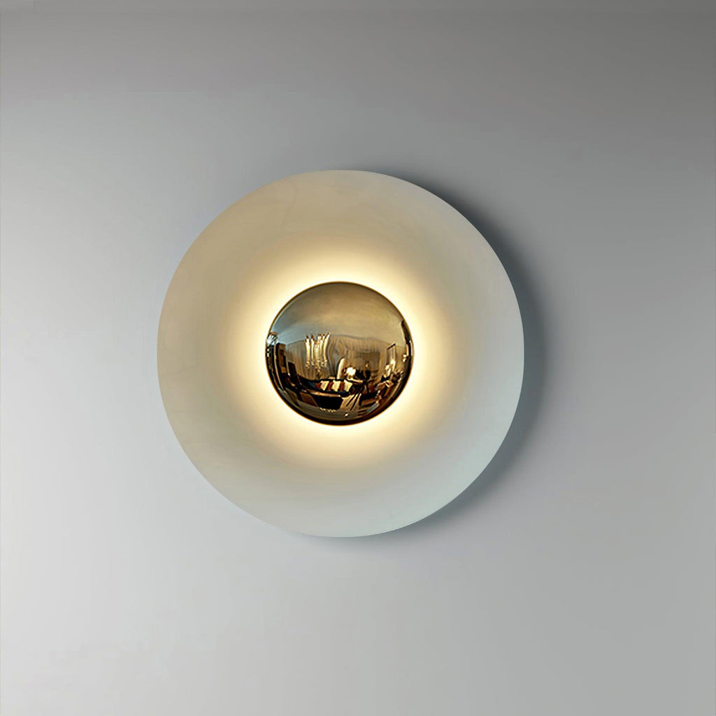 This white Clérachic wall light glows on a plain wall, its mirrored centre reflecting the room around it.