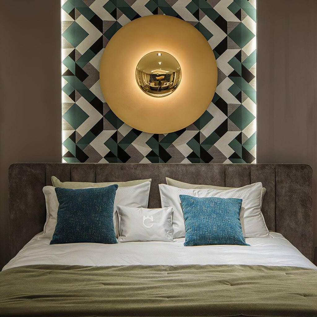 A brass-toned Clérachic wall light glows above a bed, its mirrored centre set against a geometric patterned panel on the wall.