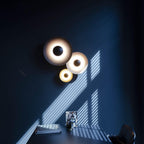 Three Clérachic wall lights glow on a dark blue wall, sunlight striping across the room from a window.