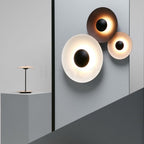 Three of this Clérachic wall light grouped on a wall, one glowing white and two dark, beside a plinth with a table lamp.