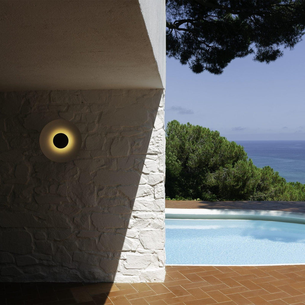 A single Clérachic wall light glows warm on a white stucco pillar, overlooking a pool and pine trees.