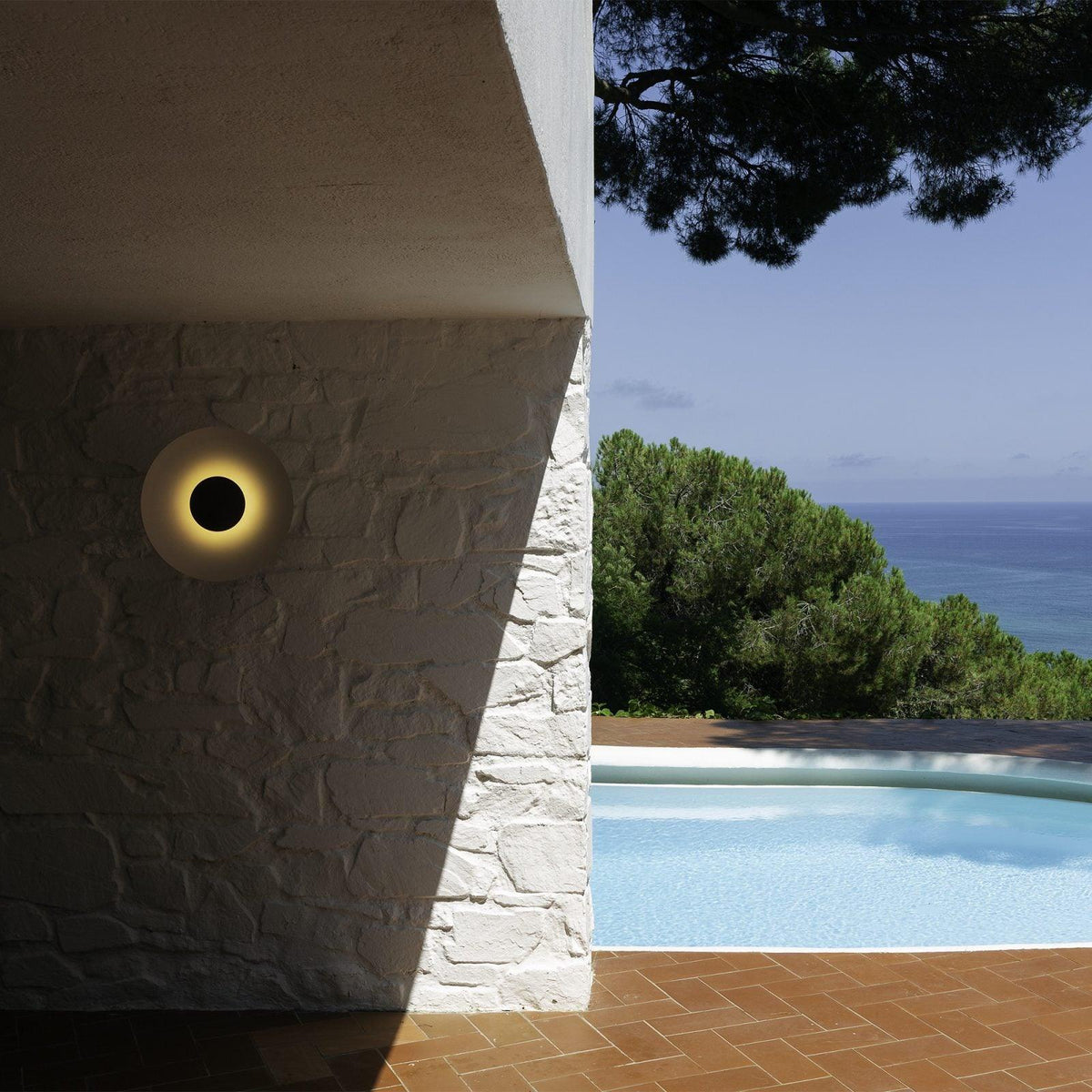 A single Clérachic wall light glows warm on a white stucco pillar, overlooking a pool and pine trees.