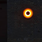 This Clérachic wall light glows bright against a dark brick wall at night, its black centre ringed with light.