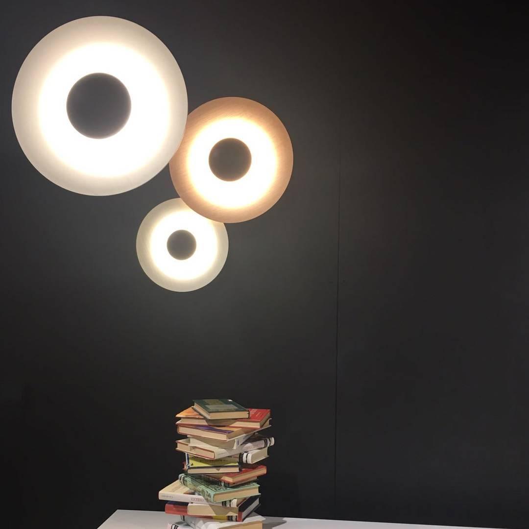 Three Clérachic wall lights glow warm on a black wall above a stack of books on a table.