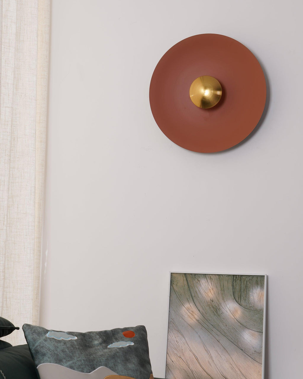 An unlit terracotta wall light from Clérachic on a plain wall, its brass centre beside a cushion and framed print.