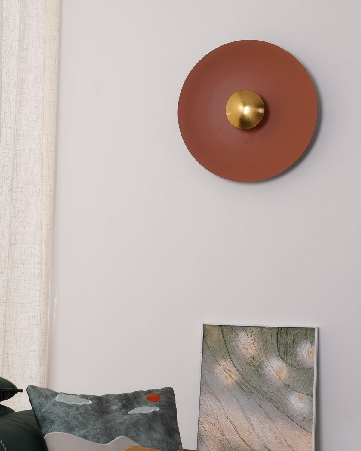 An unlit terracotta wall light from Clérachic on a plain wall, its brass centre beside a cushion and framed print.