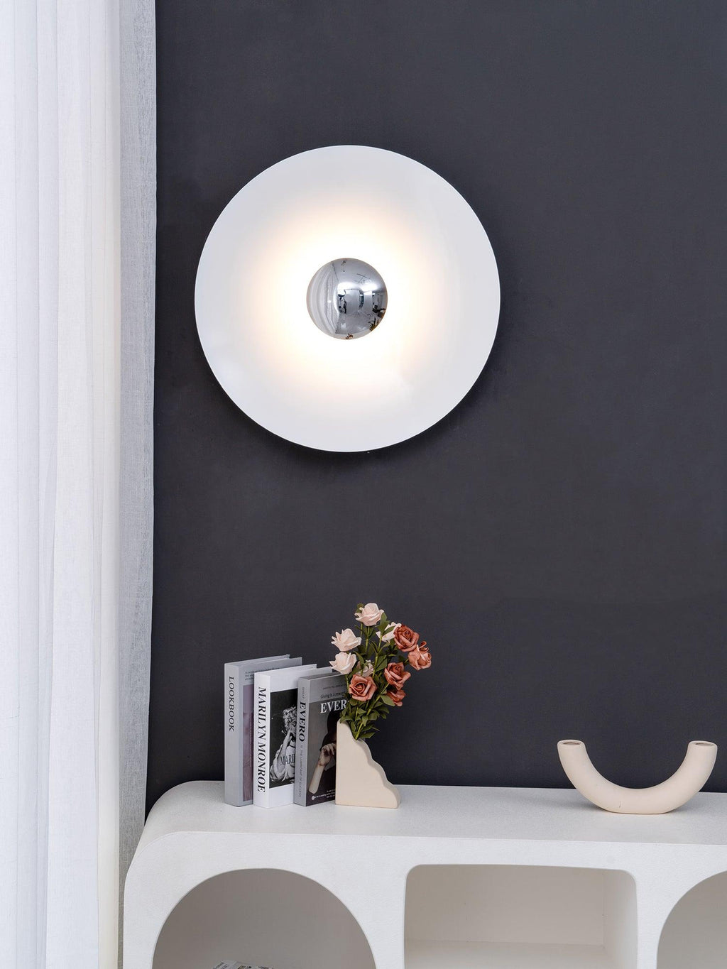 A white Clérachic wall light glows above a console table with books and flowers, against a charcoal wall.