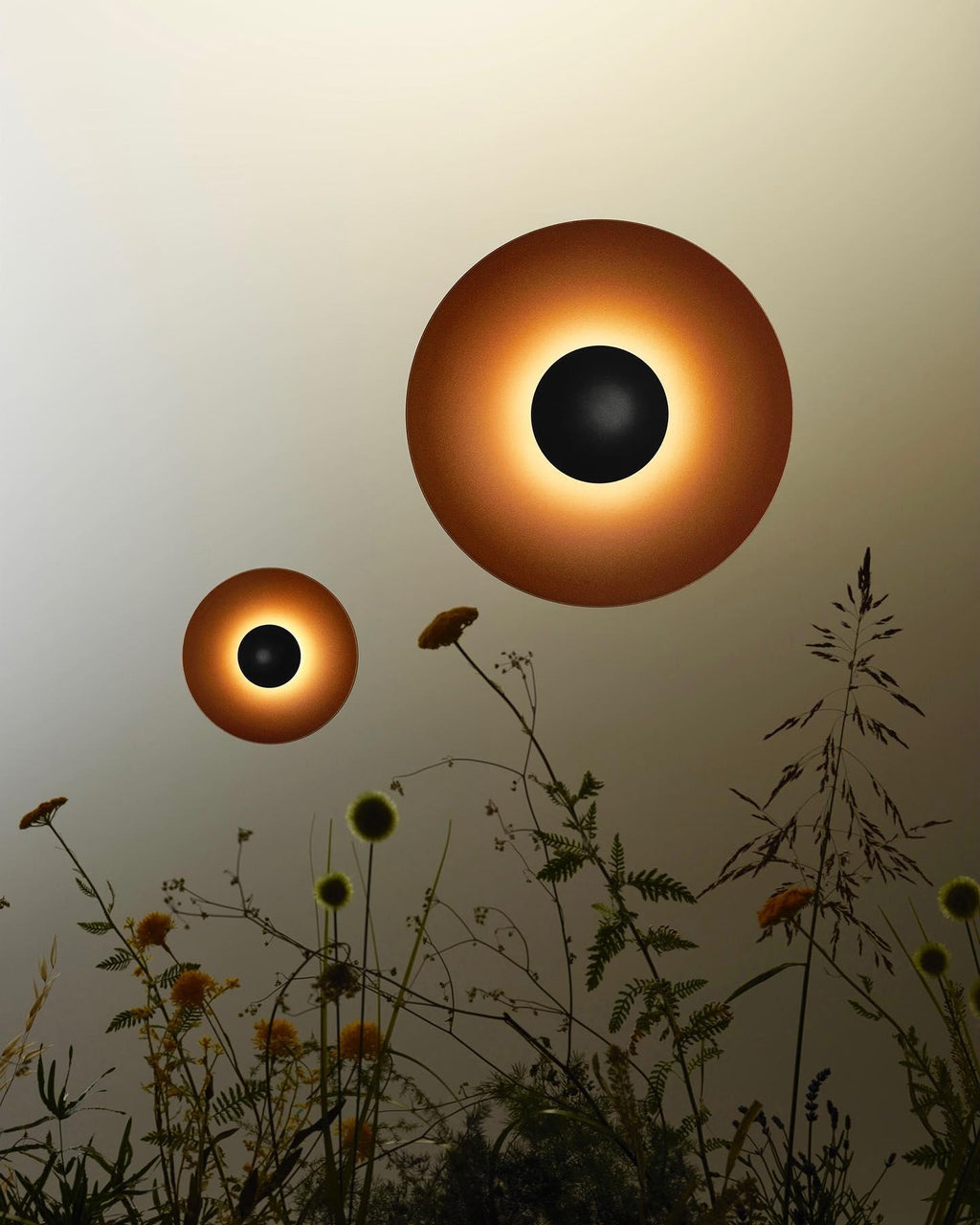 Two Clérachic wall lights of different sizes glow warm above a bed of wildflowers, shown as a styled outdoor composition.