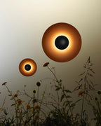 Two Clérachic wall lights of different sizes glow warm above a bed of wildflowers, shown as a styled outdoor composition.