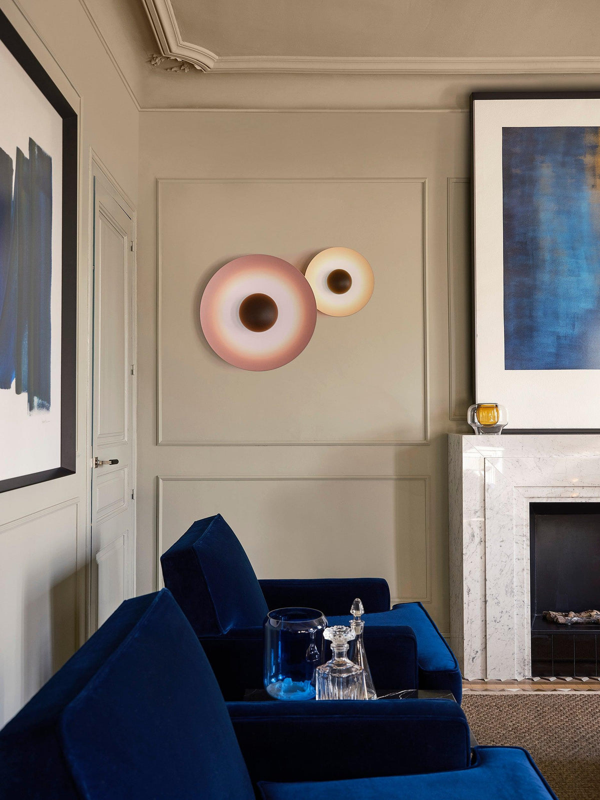 A dusky pink Clérachic wall light paired with a smaller white one on a panelled wall, near a blue armchair.