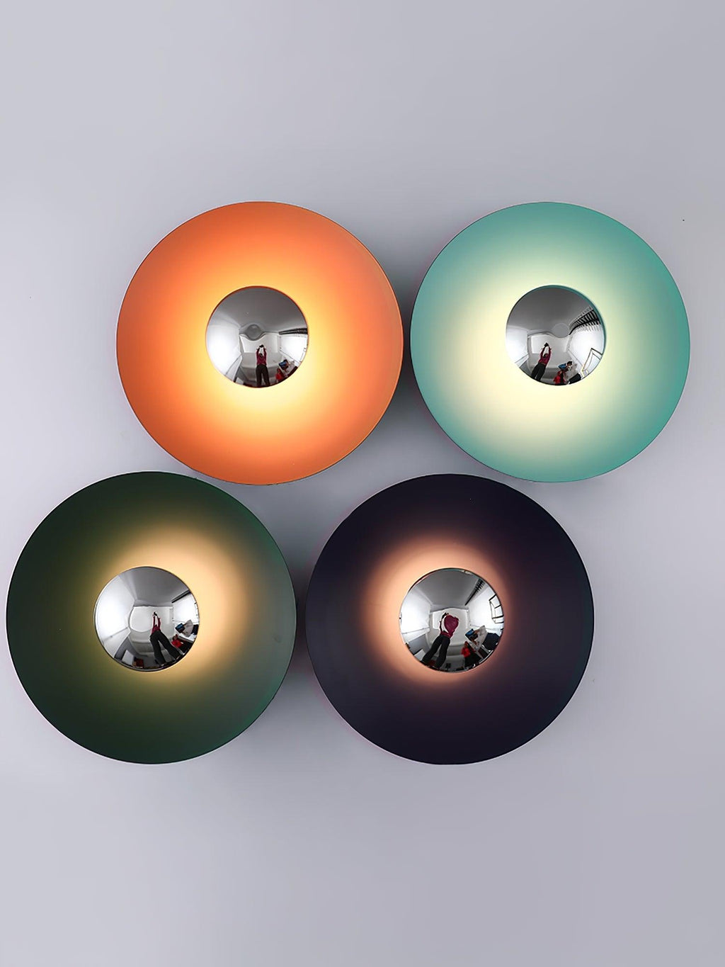 Four Clérachic wall lights in orange, mint, dark green and charcoal, each with a mirrored centre, on a plain backdrop.
