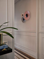 Two Clérachic wall lights, a larger pink one and a smaller rose one, on a panelled wall beside a potted plant.