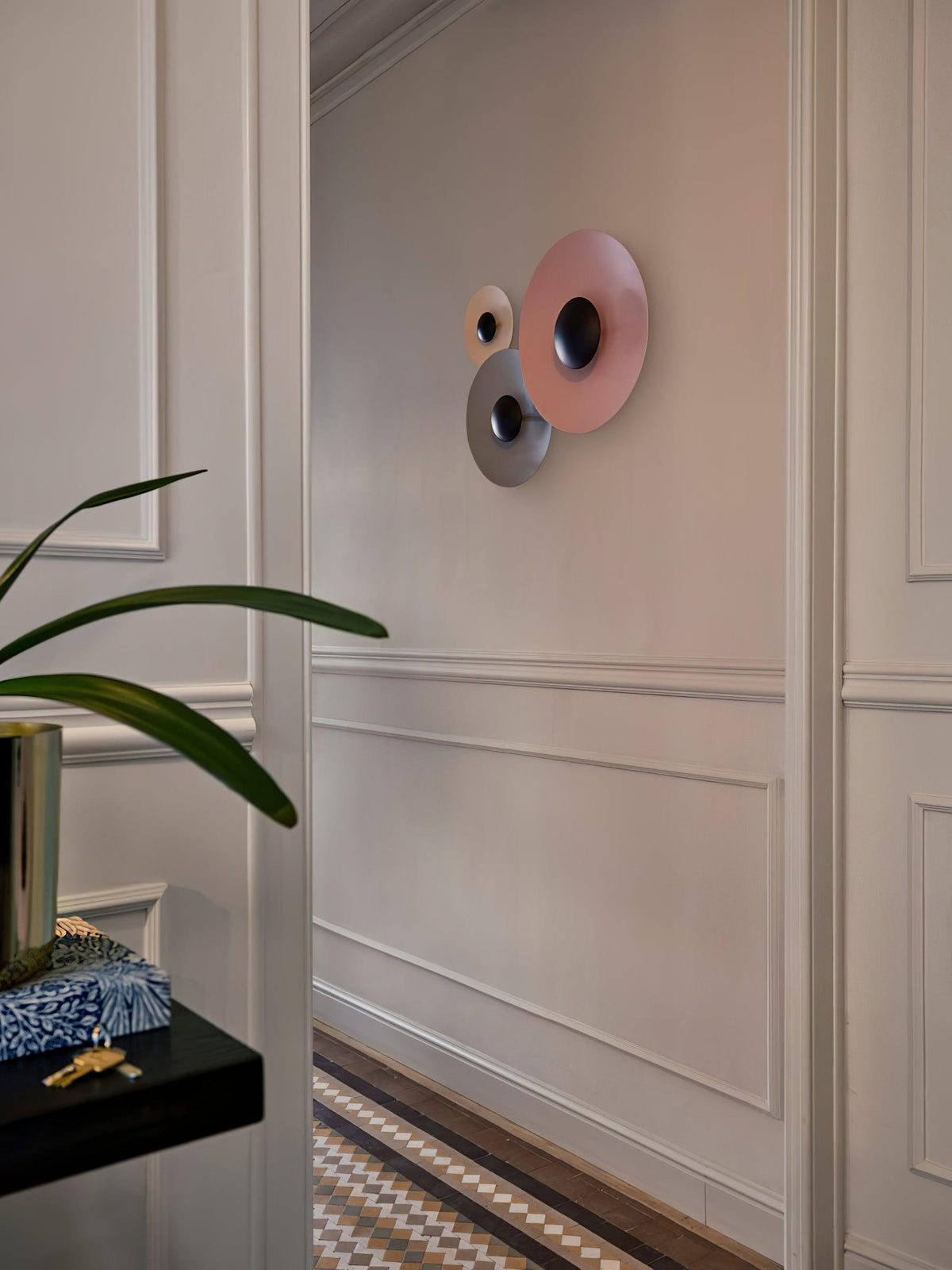 Two Clérachic wall lights, a larger pink one and a smaller rose one, on a panelled wall beside a potted plant.