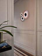 Two Clérachic wall lights in pale pink tones, grouped on a panelled wall near a potted plant and console.