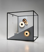 Three Clérachic wall lights glow warm inside a black wireframe cube display, beside a small pendant lamp.