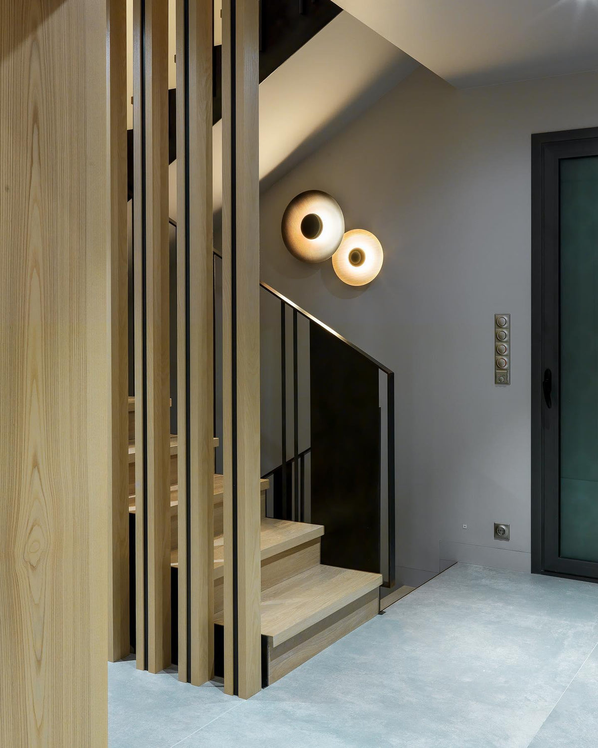 Two white Clérachic wall lights glow warm on a grey wall beside a wood-slatted staircase divider.
