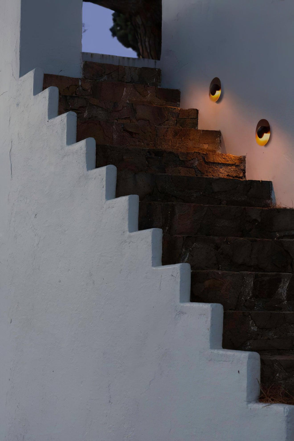 A single Clérachic wall light glows warm on a stone stairwell wall, set against a night sky.