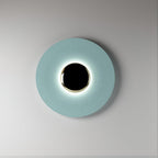 A pale blue Clérachic wall light on a plain grey backdrop, its black centre ringed by a soft glow.