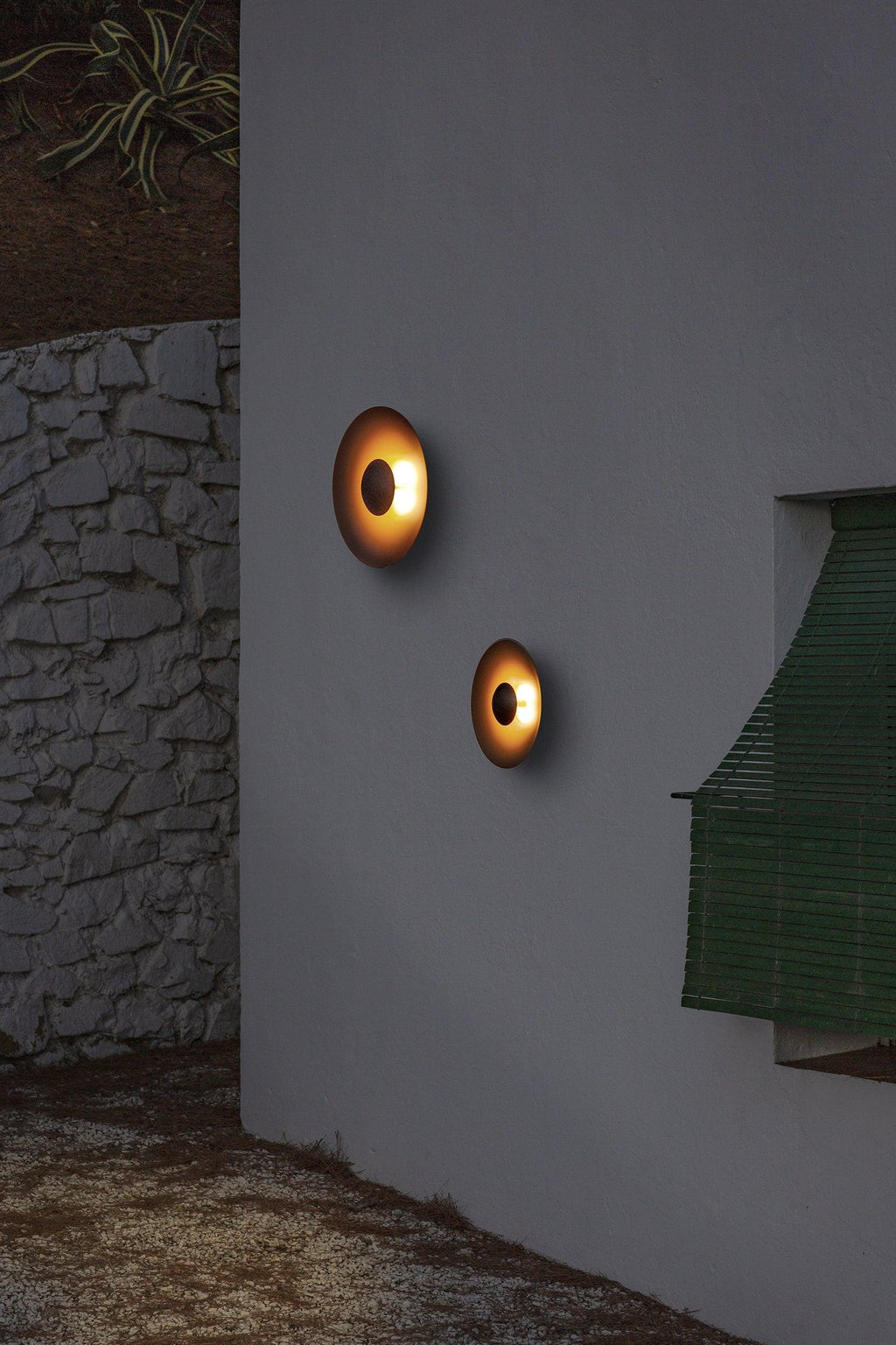 Two small Clérachic wall lights glow warm on an outdoor stone wall at dusk, beside greenery.