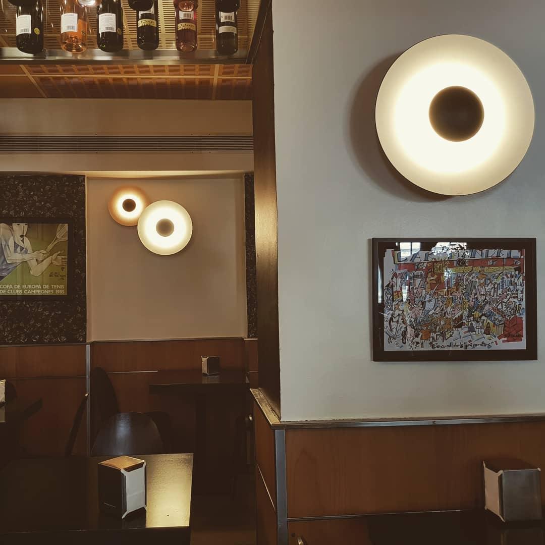 This white Clérachic wall light glows warm in a wood-panelled bar, near a rounded artwork on the wall.