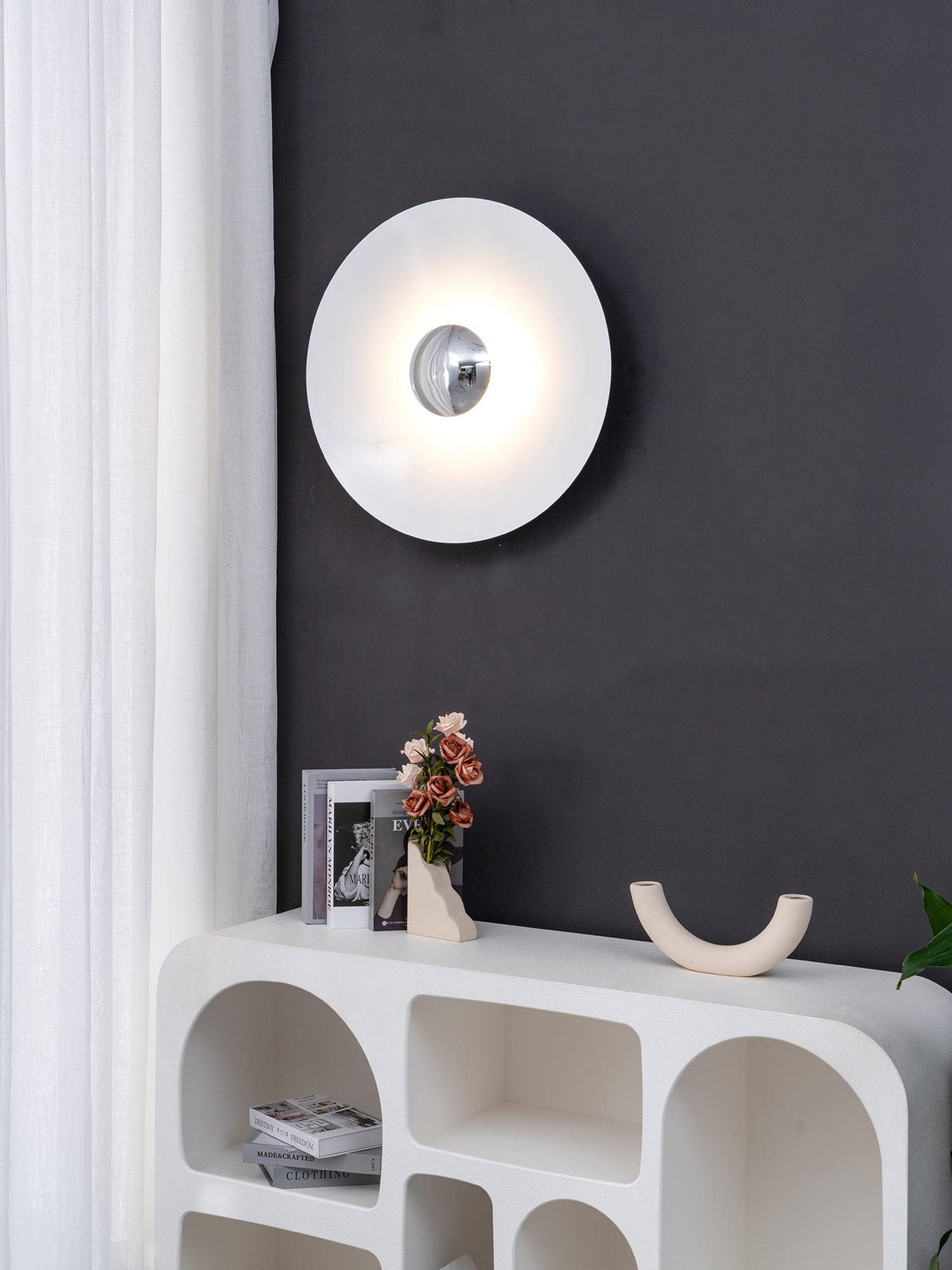 Closer view of the white Clérachic wall light glowing above a console table with books, on a charcoal wall.
