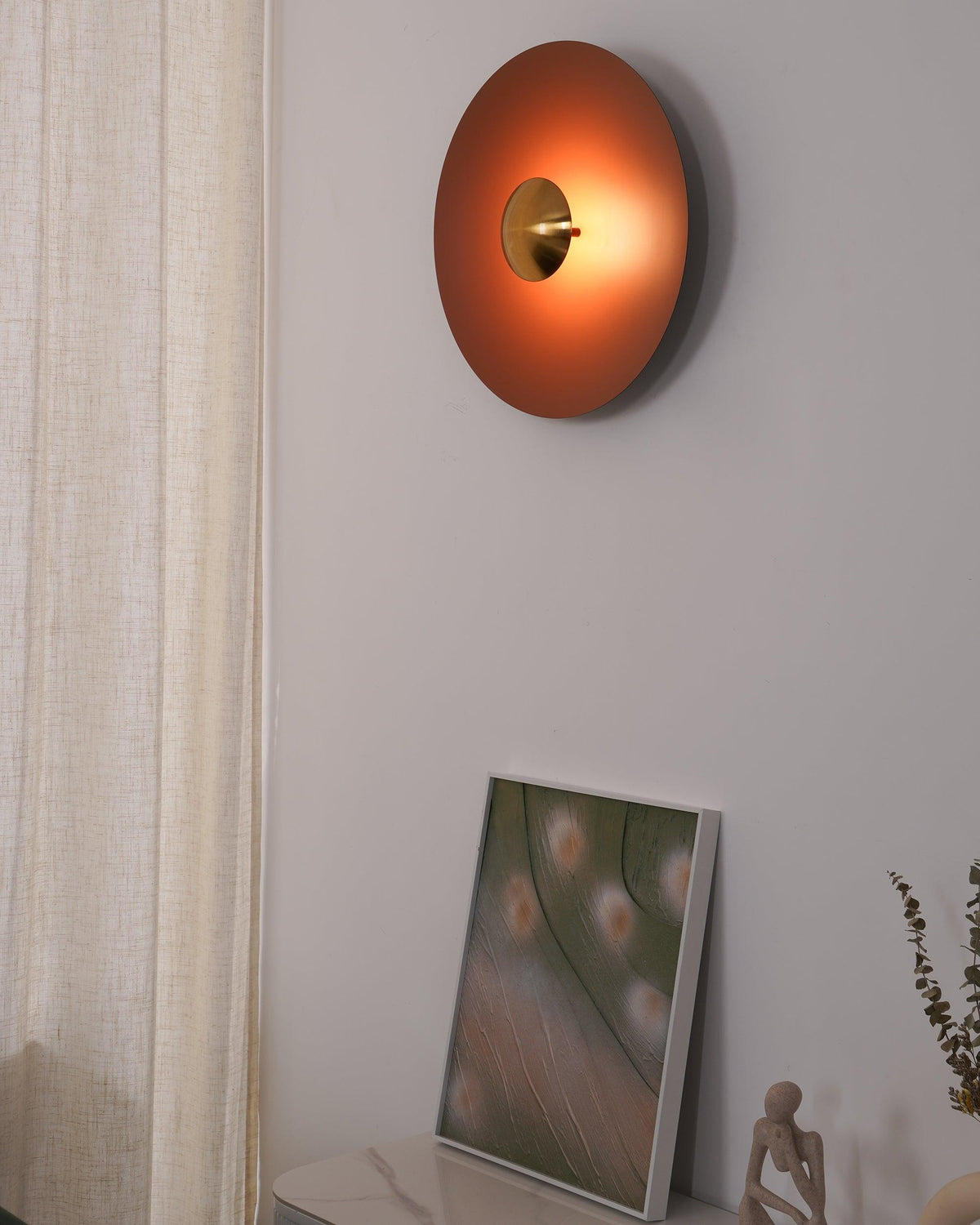 An unlit orange Clérachic wall light with an antique copper centre, near a sheer curtain and framed print.