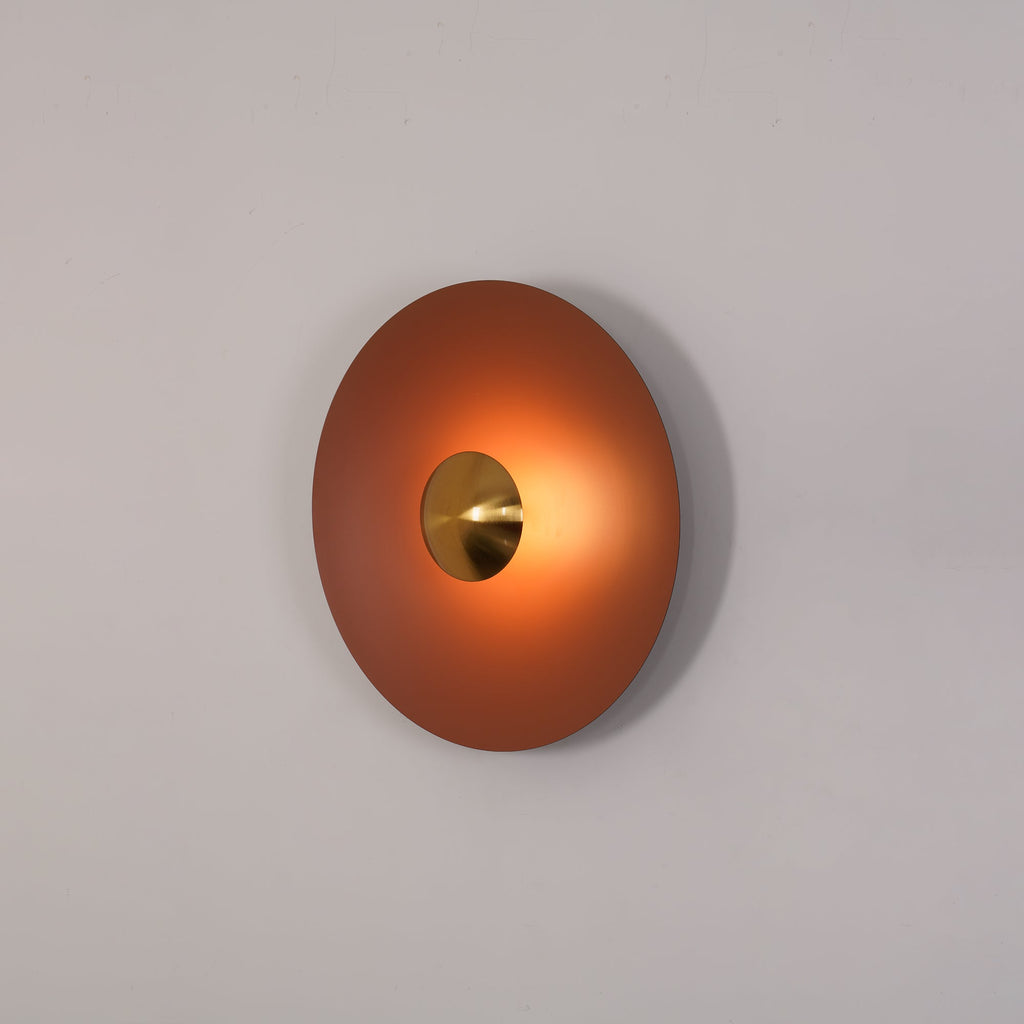 Angled view of the unlit terracotta Clérachic wall light on a plain grey wall, its brass centre visible.