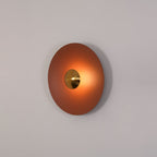 Angled view of the unlit terracotta Clérachic wall light on a plain grey wall, its brass centre visible.