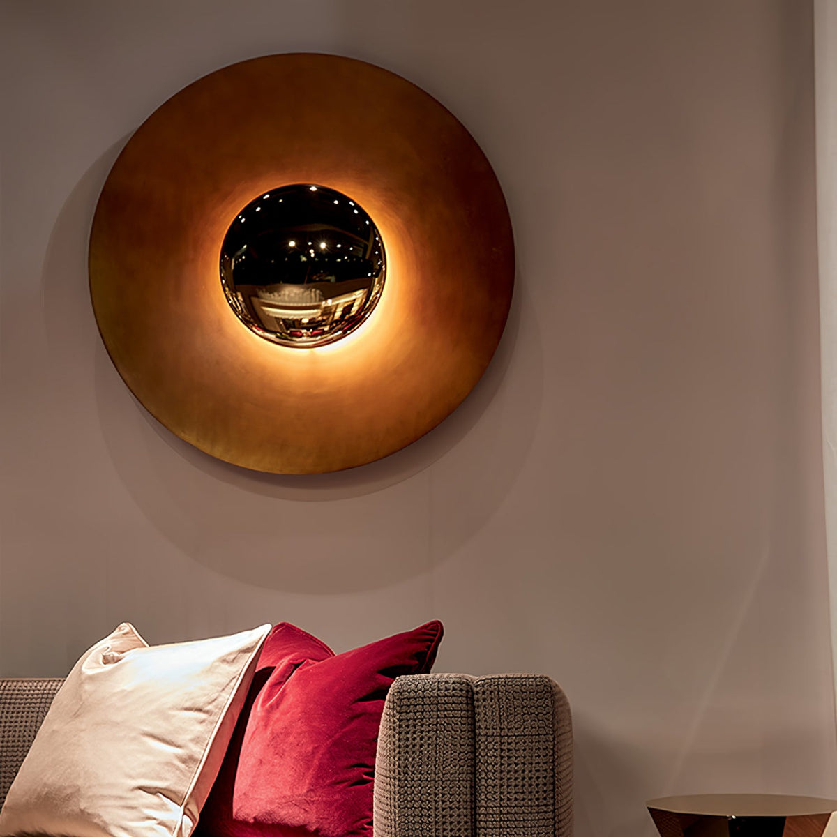 A large bronze-toned Clérachic wall light glows above a sofa with cushions, its mirrored centre reflecting the room.