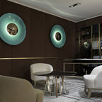 Two mint-toned Clérachic wall lights on a brown wall, one glowing warm, in a lounge with a round dark table.