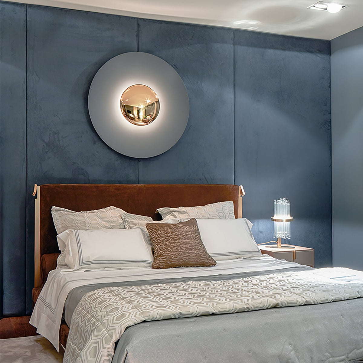 A pale grey Clérachic wall light glows above a bed with a brown leather headboard, on a dark blue panelled wall.
