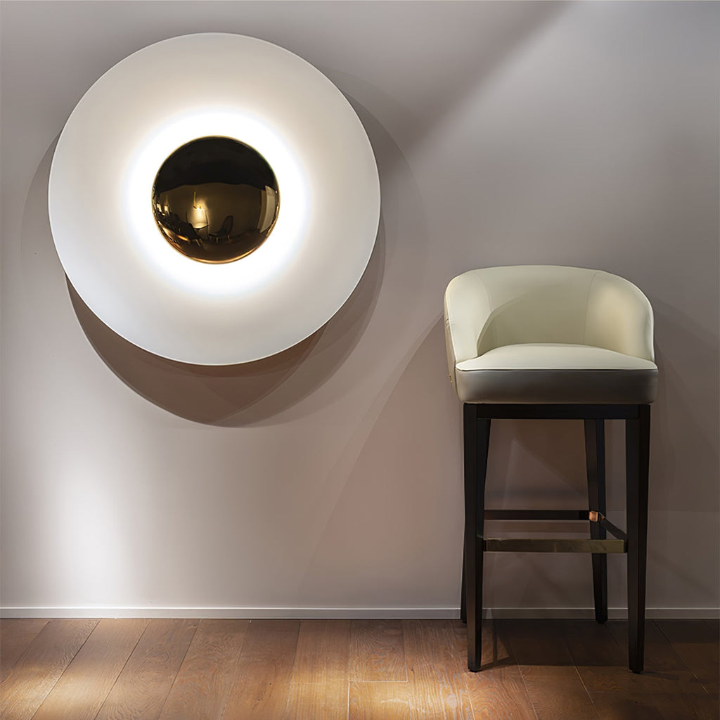 A white Clérachic wall light glows softly near a cream barstool, its brass centre warm against the pale shade.